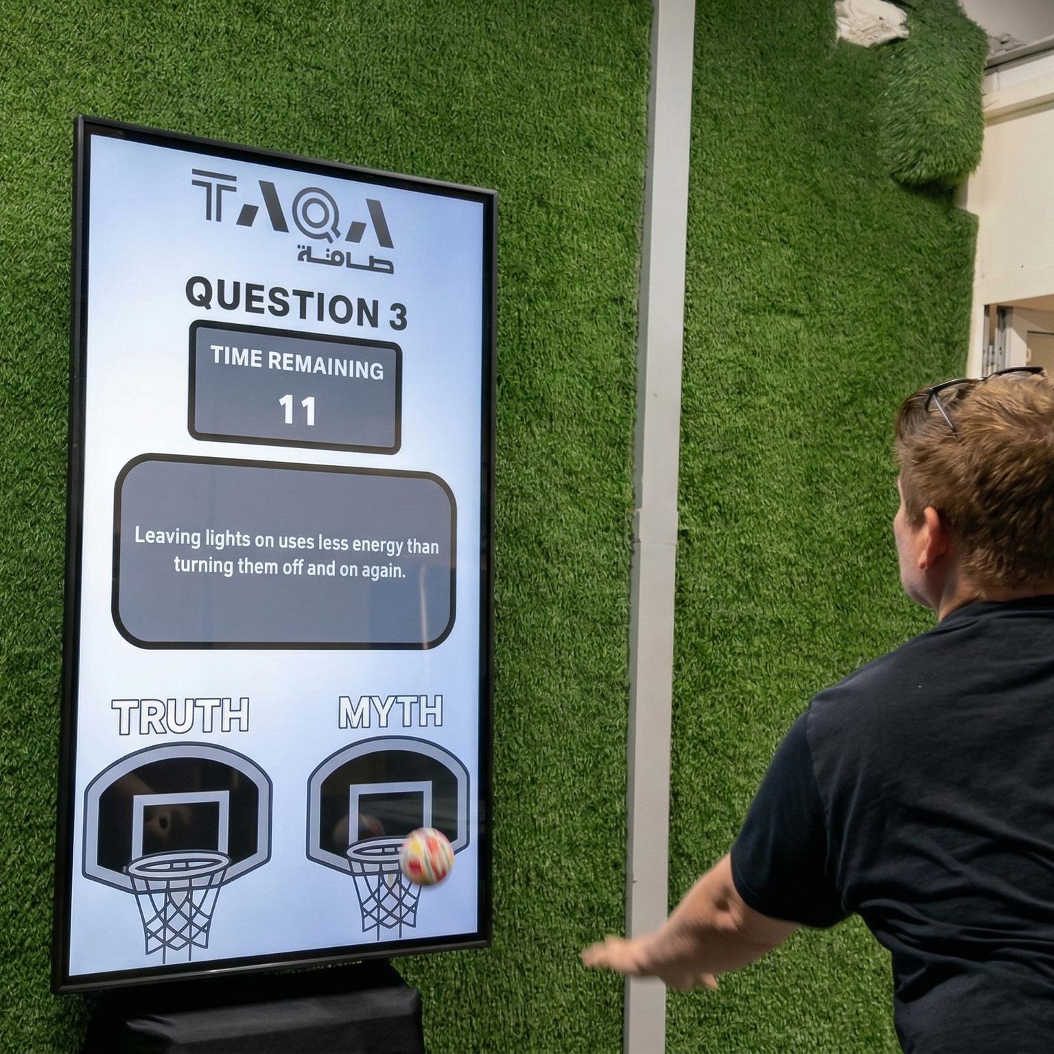 A guest throwing a softball at a large digital Truth or Myth display screen to lock in an answer at a high-energy corporate exhibition booth activation